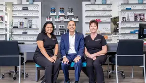 Three people, two women and a man, are sitting on a bench inside an optical shop, smiling and posing for a picture