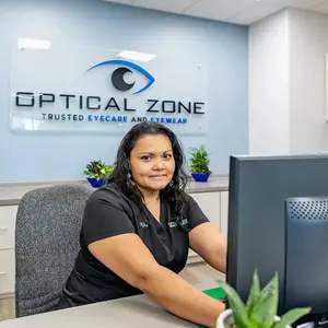A woman is sitting at a desk with a computer monitor in front of her. She is wearing a black shirt and has her hair tied up. Behind her is a wall with a sign that reads Optical Zone. On the desk are potted plants, a gray chair, and a monitor. The woman is smiling and looking at the camera.