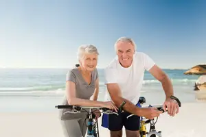 An elderly couple is riding their bicycles on the beach with a smiling face