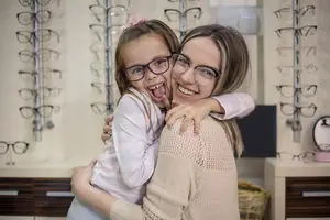 A woman and a girl wearing glasses and smiling while hugging in a shop with shelves of glasses in the background