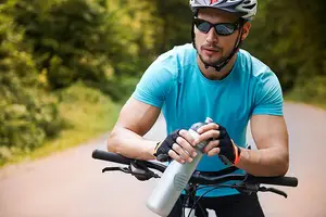 A man riding a bicycle while holding a water bottle and wearing a helmet and sunglasses.