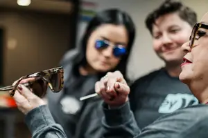 A woman holds a pair of sunglasses while another woman with blue glasses looks on and a man smiles