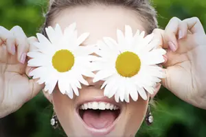 A smiling woman holds up two white daisies with yellow centers in front of her eyes, perhaps for a photo or to play a game.