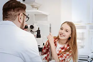 An eye doctor uses a microscope to examine a young girl's eyes while she smiles.