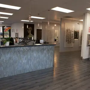 An interior view of an optometrist's office with a reception desk, framed pictures, and various decorative elements, including a picture of a woman wearing glasses and a potted plant, and a rack of eyeglasses on the wall.