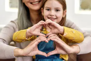 A smiling woman and a young girl are making a heart shape with their hands, posing for a photo.