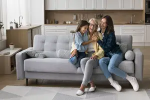 Three women are sitting on a gray couch in a modern kitchen with white cabinets, a wooden counter, and a sink with a faucet.