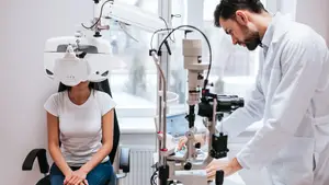 A man in a white lab coat adjusting an eye exam machine on a woman sitting in a chair in front of a window