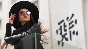 A woman wearing a black hat and sunglasses poses for a photo in front of a building with Asian characters on the wall.