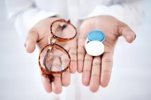 A person wearing a white long-sleeved shirt is holding a pair of glasses and a blue-lidded container in their hands. The glasses have a brown frame and clear lenses. The container is likely a lens case. The person's hands are slightly blurry, and the background is white.