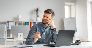 A man is sitting at a desk in an office holding his head in his hands.