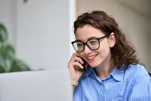 A woman wearing glasses and a blue shirt with white stripes is talking on her cell phone while sitting in front of a laptop on a desk.