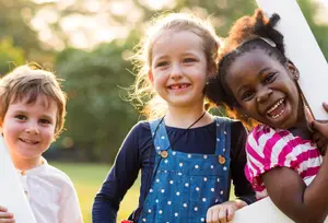 Three kids posing for a picture in a park, the girl on the left is holding a white object, and the girl in the middle is wearing a denim jumper with white polka dots.