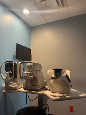 A room with medical equipment, including an eye examination machine, a monitor, and a stool, is illuminated by a ceiling light, with a power outlet on the wall.
