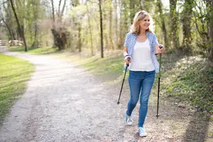 A woman is walking on a pathway with a smile on her face and holding trekking poles