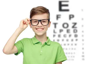 A young boy wearing glasses is smiling and holding his glasses in front of an eye chart.