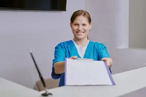 A woman wearing a blue lab coat smiles at the camera as she stands behind a reception desk.