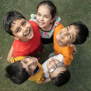 Five young children, all wearing different colored shirts, are standing in a circle on a grassy field, smiling and posing for a photograph. The girl in the center is wearing a floral shirt, and the boy on the left is wearing a red shirt, while the boy on the right is wearing a white shirt with glasses.
