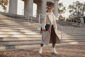 Woman wearing glasses, a scarf, a white coat, and white boots standing on brick pavement with a smile on her face while holding a folder.