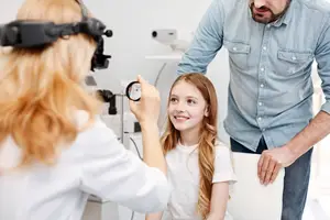 A young girl is having her eye examined by an adult female wearing a stethoscope and a man standing behind her in an indoor setting.