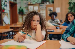 A girl with curly hair is sitting in a classroom, holding a pen, and looking at something on her desk.