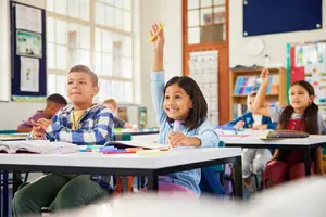Three children sitting at a desk in a classroom, one raising their hand to ask a question