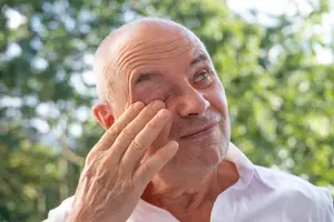 An older man with wrinkles on his face, wearing a white polo shirt, touches his right eye with his left hand, probably in a park or garden.