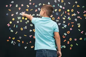 A young boy is standing in front of a blackboard with colorful alphabets and numbers arranged in a creative pattern, he is pointing at a number with his left hand.