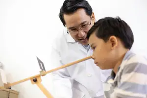 An adult male doctor wearing a white lab coat is examining a young boy with a medical instrument
