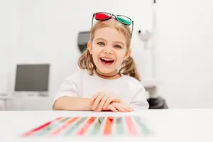 A little girl wearing 3D glasses is sitting on a chair and smiling, possibly in an optical clinic. There is a table in front of her with some colorful paper strips, and a monitor is on the desk behind her. The room has white walls and a blurry background.