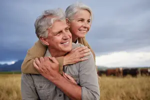 A senior man and woman are smiling and hugging each other in a field with cows.