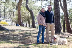A man and a woman are standing in a park with a dog, both smiling and looking at the camera. They are wearing casual clothing and the dog is lying on the ground. There are trees and a building in the background, and it seems to be a sunny day.
