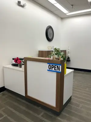 A reception desk with a white countertop and a brown wooden base with a sign that reads OPEN. There is a flower vase on the countertop, a potted plant on the right side, and a clock on the wall above the desk. On the left side, there is a black luggage with a flower on it. The room has a white wall, a ceiling light, and a tiled floor.