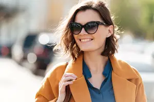 A smiling woman wearing sunglasses and a brown coat on a street