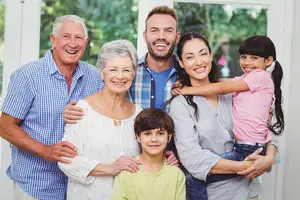 A smiling family of five, including an elderly man, a woman, a man, a boy, and a girl, posing for a photo in a room with a glass window and greenery outside