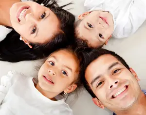 A smiling family of four lying on a white surface, likely in a room, with a child in the center looking directly at the camera