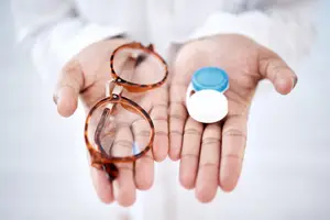 A person is holding a pair of glasses and a contact lens container in their hands, standing in front of a white wall. The person is wearing a white long-sleeve shirt and is holding the glasses and contact lens container in their hands, possibly in a laboratory setting.