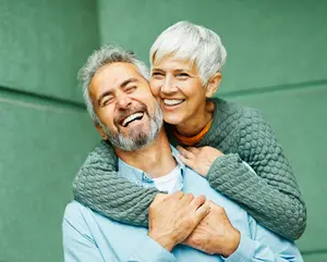 A smiling elderly couple hugging and posing for a photo in front of a green wall