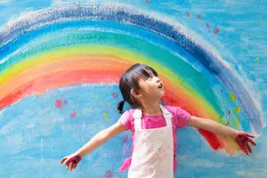 A young girl with pink paint on her hands is standing in front of a colorful rainbow mural