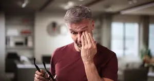 Image of an older man with gray hair wearing a maroon t-shirt holding glasses while rubbing his eye with his hand in an indoor area