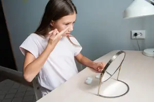 A young girl is examining her face in a mirror while holding her nose.
