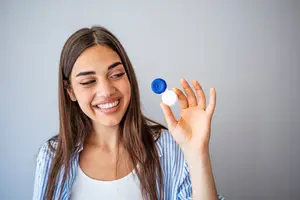 A smiling woman holding a contact lens case with a blue lid and white base in her hand, wearing a blue and white striped shirt, standing in front of a gray background. The contact lens case is in the shape of a pill, and the woman is looking at it with a smile. She has long, straight hair and is wearing eyeliner.