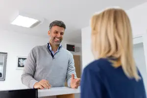 A man in a gray sweater and a woman with blonde hair standing near a white desk and talking