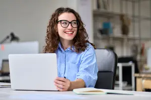 A smiling woman wearing glasses is sitting in a chair in an office, holding a laptop, and surrounded by various items such as books, a monitor, a shelf, and a lamp.