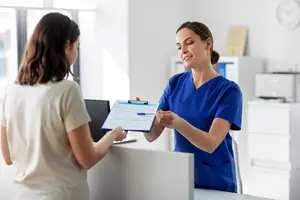 A woman in a blue uniform holds a clipboard and a pen while speaking with another woman in a white shirt in a clinic setting.