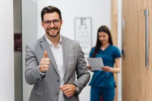 A man in a gray suit giving a thumbs up while a woman in blue scrubs walks behind him in a hallway.