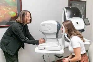 An adult woman is assisting a young woman in an eye examination at a clinic using a modern eye-checking machine.