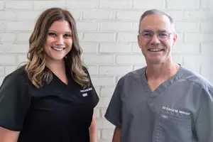 Two people smiling for a photo, a woman in black and a man in gray, in front of a white brick wall.