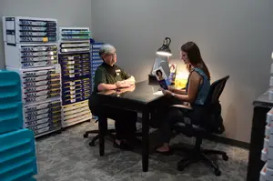 Two women sit at a desk with a lamp and paper, wearing glasses, while the woman on the right holds a paper and a pen, and the room has carpeted floors, shelves with boxes, and a desk lamp.