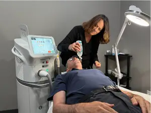 A man is lying on a bed while a woman is standing beside him holding a bottle and appears to be giving him some treatment